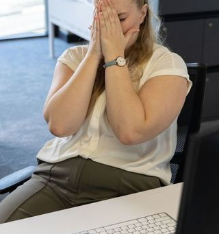 Woman at a desk covers her face with hands, appearing stressed. She's in an office with a computer keyboard nearby, wearing a watch. Suggesting burnout.