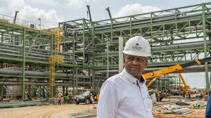 Dangote in a white shirt and helmet stands at a construction site with metal structures. Cranes and workers are visible under a cloudy sky.
