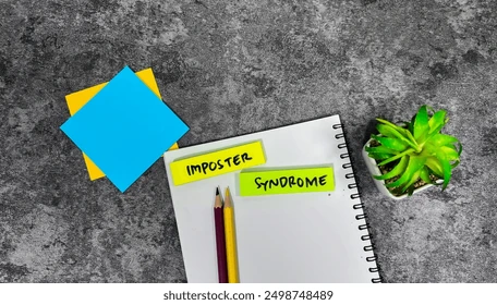 Notebook with "Imposter Syndrome" stickers, two pencils, sticky notes, and a small green plant on a gray textured surface.