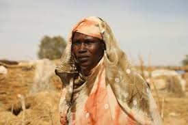 A Sudanese woman in an orange and beige patterned headscarf stands outdoors, with hay bales in the background under a clear sky. She appears thoughtful.