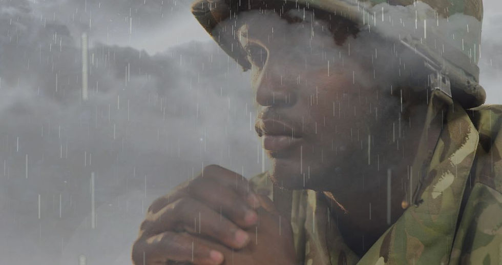 A soldier in camouflage holds hands in thought, rain falling, clouds in the background. The mood is contemplative and somber.