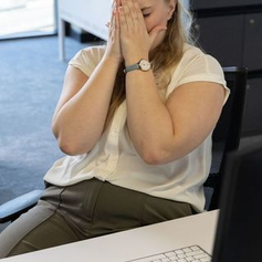 Woman in white shirt sits at desk, covering face with hands, appearing stressed. Suggesting emotional burnout.