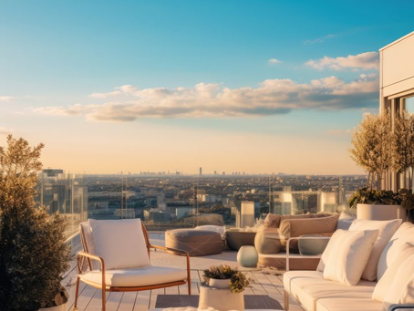 Rooftop terrace at sunset with white lounge chairs, plants in pots, and a glass railing overlooking a cityscape under a blue sky.