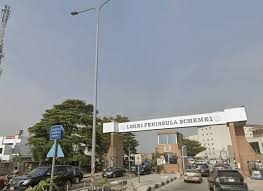 Street view of Lekki Peninsula Scheme 1 entrance. Cars pass under a white archway with visible text, surrounded by trees and buildings.