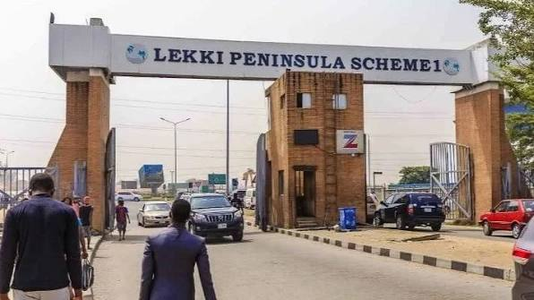 Entrance gate to Lekki Peninsula Scheme 1 with pedestrians, cars, and brick pillars under a bright sky.
