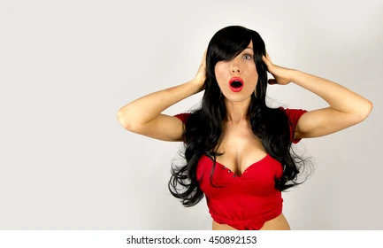 Woman in red top with long black hair, surprised expression, hands on head against gray background. 