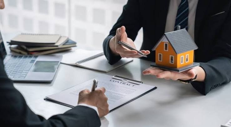 Two suited professionals sign a contract at a desk, with a small house model, laptop, and notebooks in a real estate investment office.