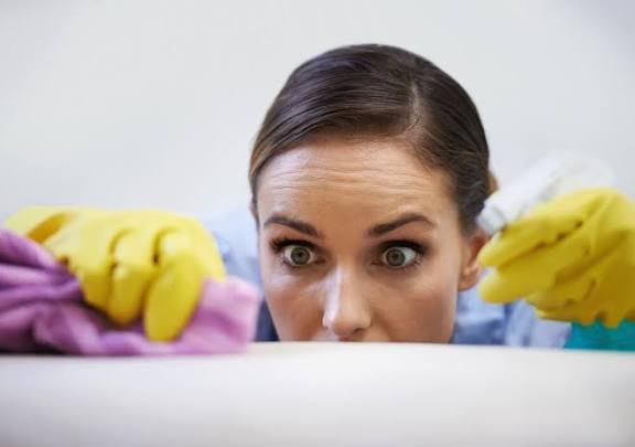 Woman peeks over a surface while colorful gloved hands clean around her, looking wide-eyed and surprised in a bright room.