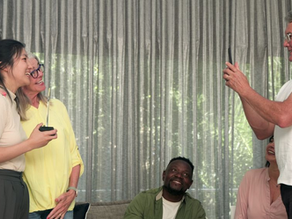 A group of people smiling as one takes a photo with a phone. Two are standing, with beige and yellow shirts, others seated. Group therapy.
