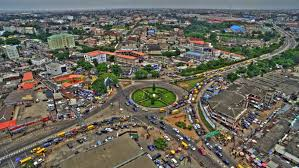 Aerial view of Ikeja surrounded by buildings and green spaces. Numerous vehicles and colorful rooftops create a vibrant scene.