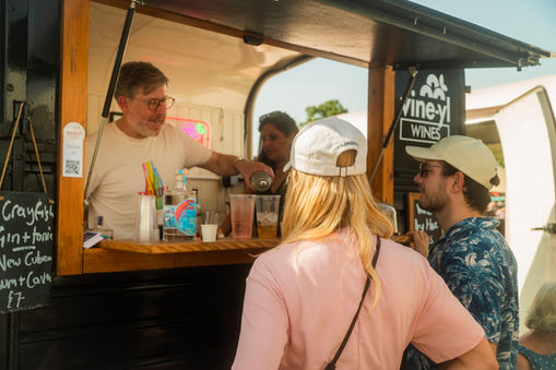 A bartender pouring a drink for customers gathered at the window of a "vineyl wines" trailer.