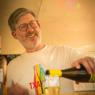 A bartender with glasses and a white t-shirt carefully pouring wine into a glass.