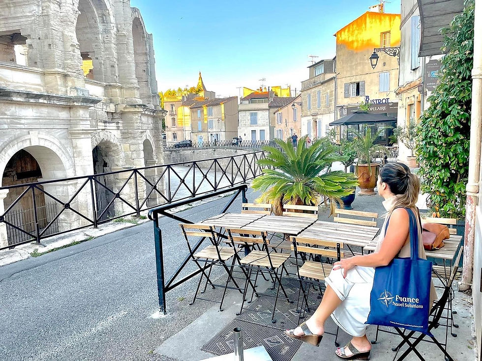 Maud Dunne with France Travel Solutions tote bag sits at outdoor café near ancient stone structure under clear sky in Arles, surrounded by potted plants and quaint buildings.