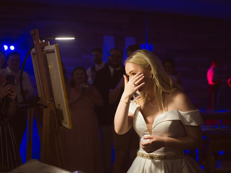 Bride in a white dress wipes tears while smiling at a painting. Guests watch in dim, colorful lighting. Emotional, intimate scene.