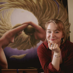 Smiling woman in red shirt poses in front of a painting of a nude figure with swirling golden hues. Warm, artistic atmosphere.
