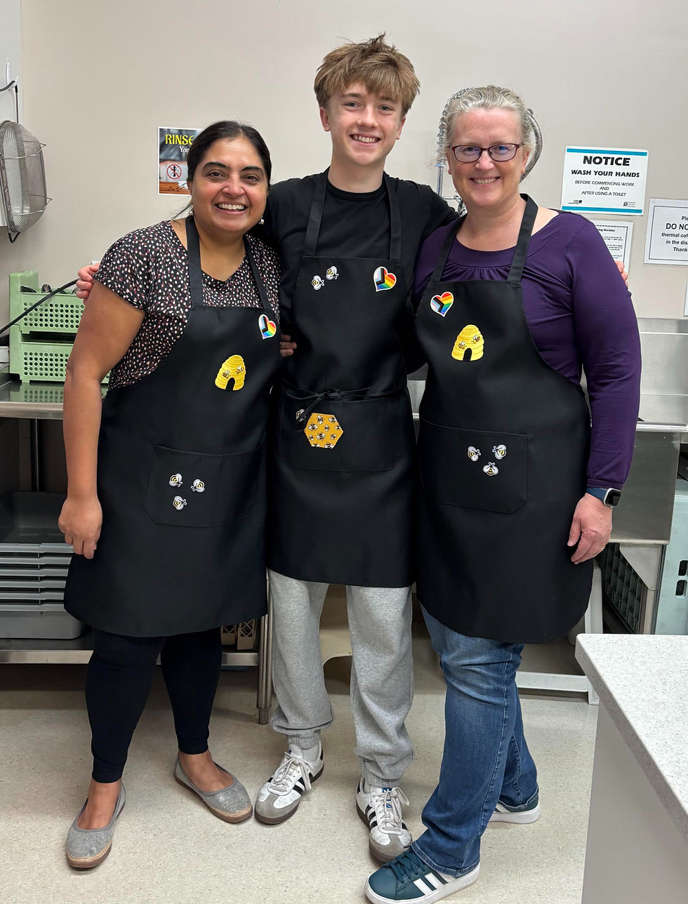 Three people wearing matching black aprons with bee and heart designs, smiling in a commercial kitchen.