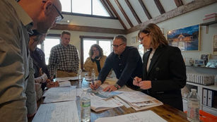 People reviewing architectural plans on a wooden table in a vaulted room. Bright window, exposed beams, and a focused, collaborative mood.