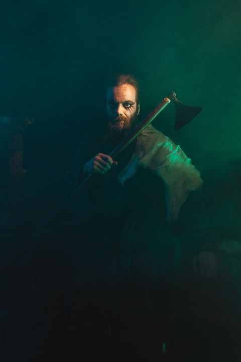 Photo de la série photographique Primitif. Sur le thème du Moyen Âge et de la fantaisie, Il est armé d'une hallebarde dans un environnement sombre, froid et vert. Il est assis sur un banc en bois avec des peau de bêtes dessus et une peau de chèvre sur l'épaule, plan moyen de face.
Il prend une pose cérémoniale de Jarl.