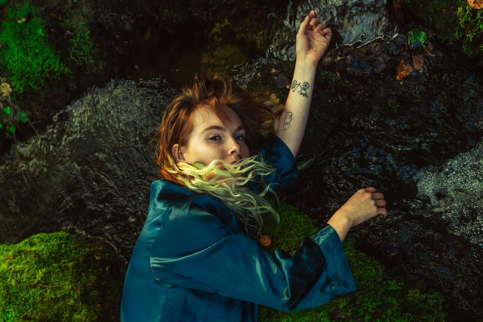 Photographie d'une femme dans une forêt allongée sur le bord d'une rivière, sur des rochers et de la mousse. Elle est vêtue d'une toile bleue.