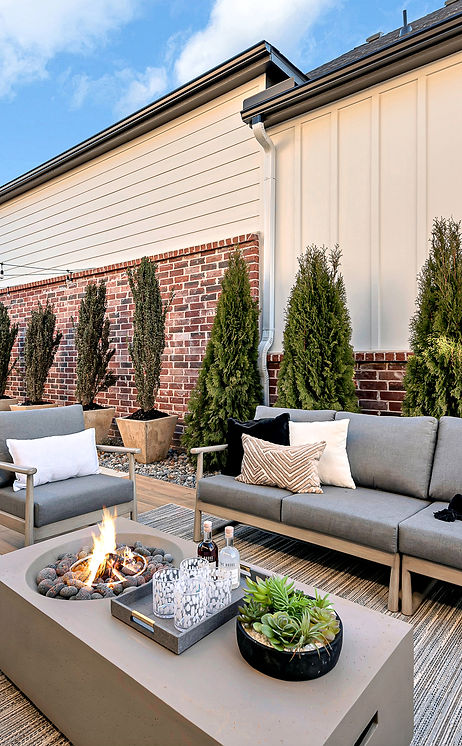 Modern patio with fire pit, gray outdoor seating, dining area, string lights, and privacy trees against brick and siding wall