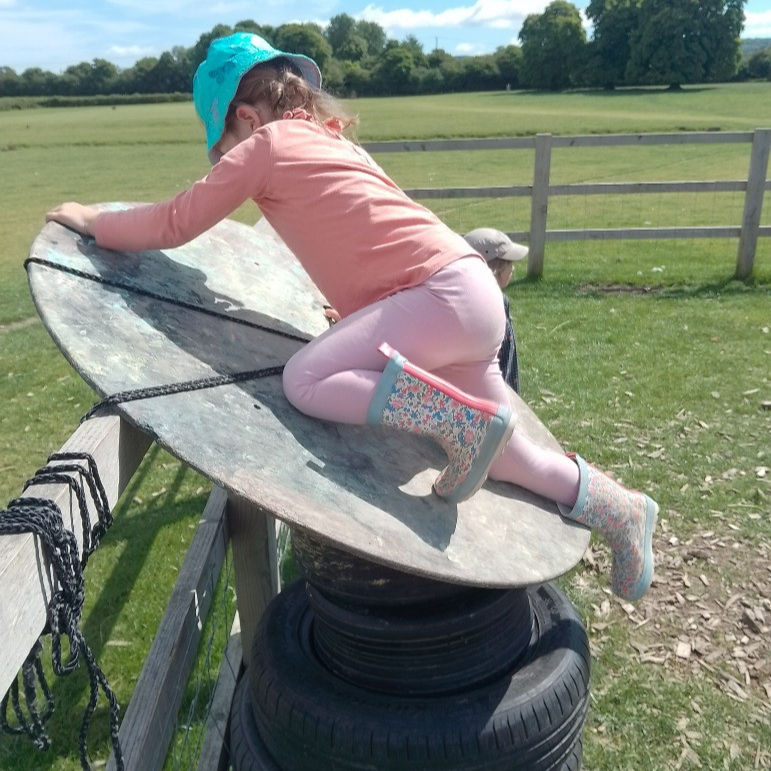 A girl wearing wellies and a sun hat is balancing on a wooden circle, which is balancing on a tower of tyres leaning against a fence. She is in a field.
