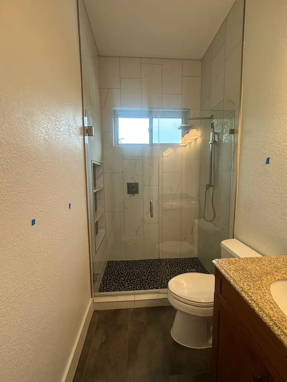 Modern bathroom remodel with frameless glass shower, large marble-look wall tile, black penny shower floor tile, recessed shower niche shelving, dark wood-look flooring, and wood vanity with granite countertop in a contemporary Austin home.