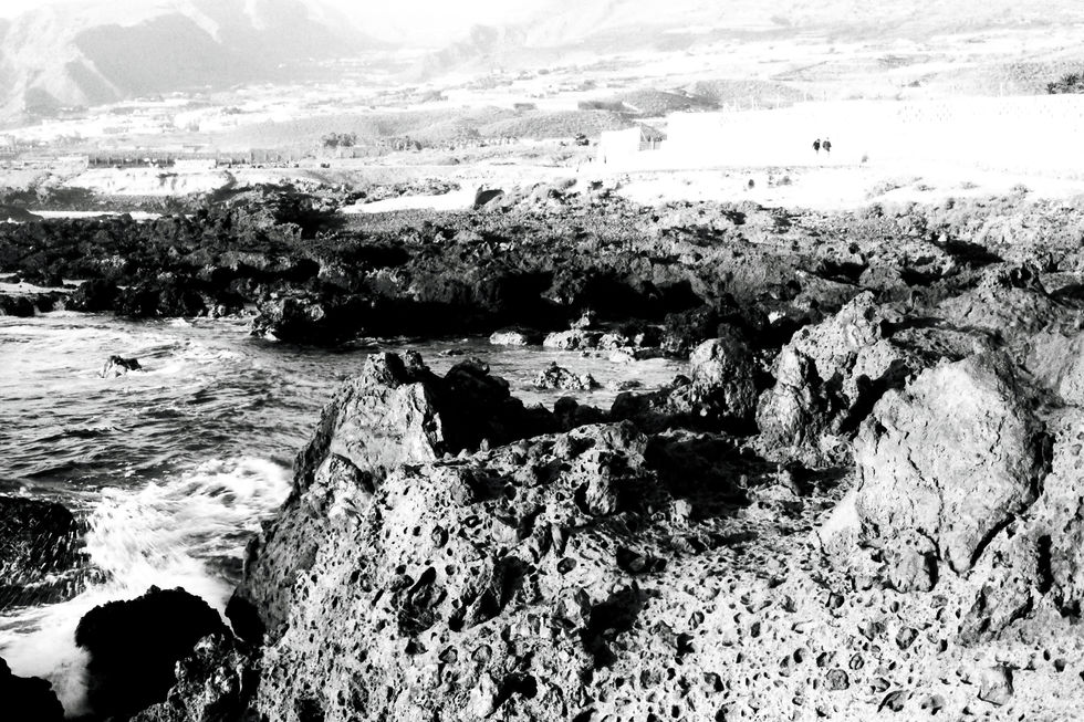 Volcanic coastal landscape in Tenerife with eroded lava rock formations and porous textures. Waves crashing against black volcanic rocks, high-contrast black and white analog photography.