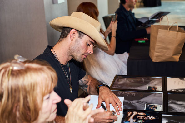Male actor sitting at a table looking down signing autograph.