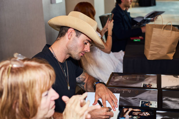 Male actor sitting at a table looking down signing autograph.