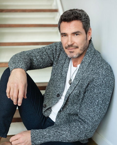 Man with short beard wearing grey cardigan sitting on wooden stairs.