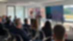 Business presentation in a bright office; woman speaks to seated audience beside screen reading A Welcoming Home to Global Trade.