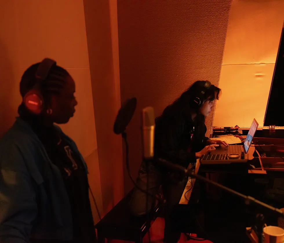 Two women wearing headphones in a music studio. One is seated with a laptop, while the other is standing in front of a microphone. Warm lighting, cozy and collaborative atmosphere.