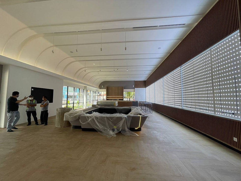 Three people discussing in a modern room with wooden floors, large windows, and covered furniture. Bright LED panel lights line one wall.