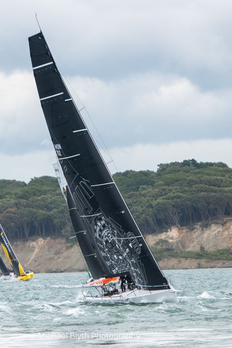 Admirals Cup 2025 Jolt 3 with black sail racing on choppy waters, showcasing white design and number "Mon 52". Green trees and cloudy sky in background.