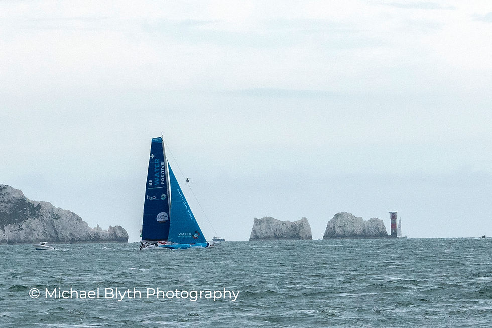 Fastnet race 2025, yacht Emira VI with "BE WATER POSITIVE" text sails near close to The Needles Lighthouse on a cloudy day. Wavy sea, distant boats visible.