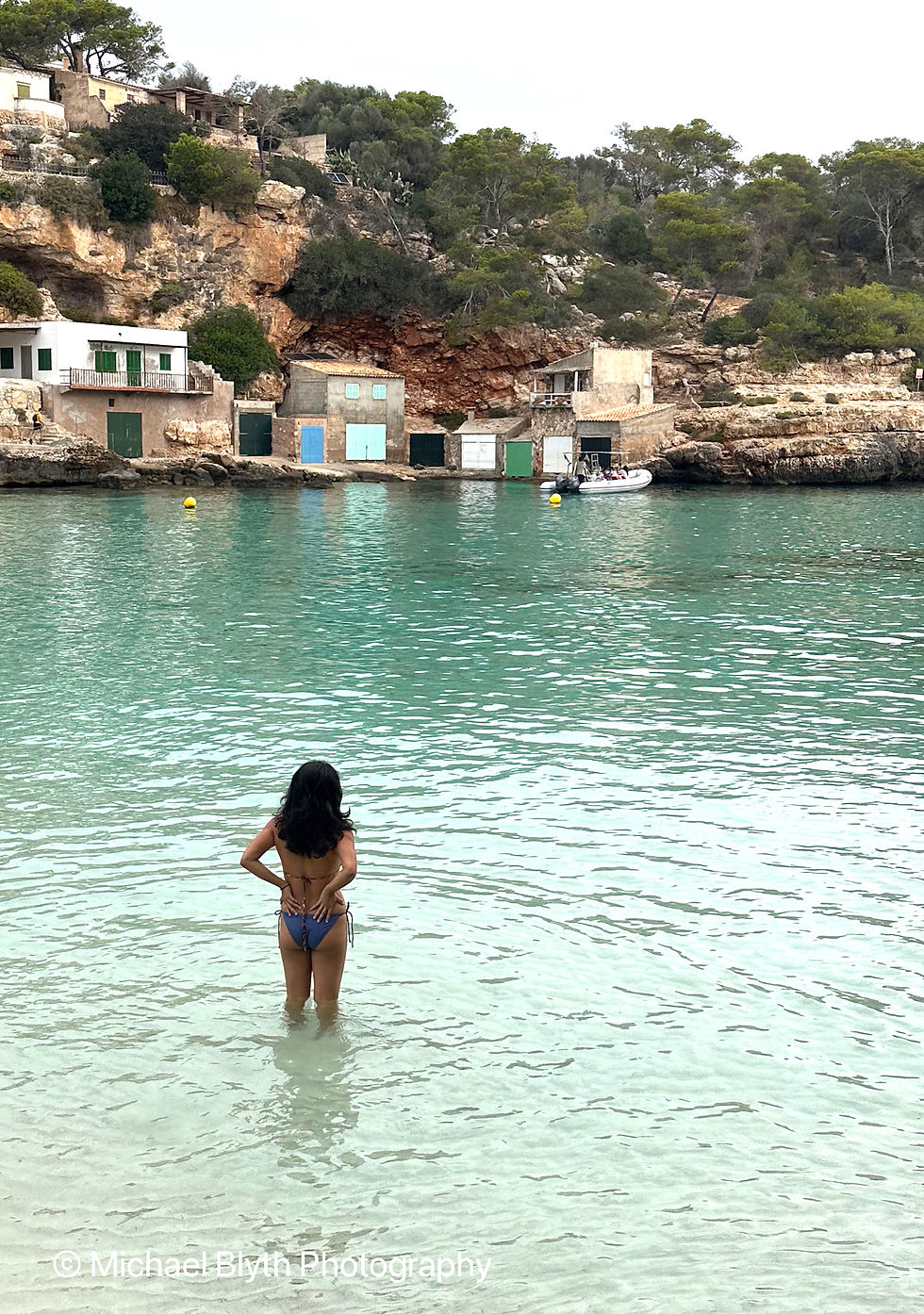 Woman in bikini walking in turquoise sea at Cala Llombards with limestone cliffs behind her.