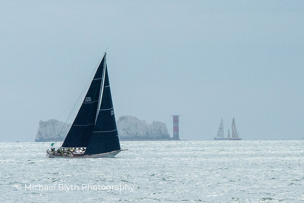 Fastnet race 2025, yacht Hound with a dark sail navigates calm waters near distant cliffs and a striped lighthouse under a cloudy sky.