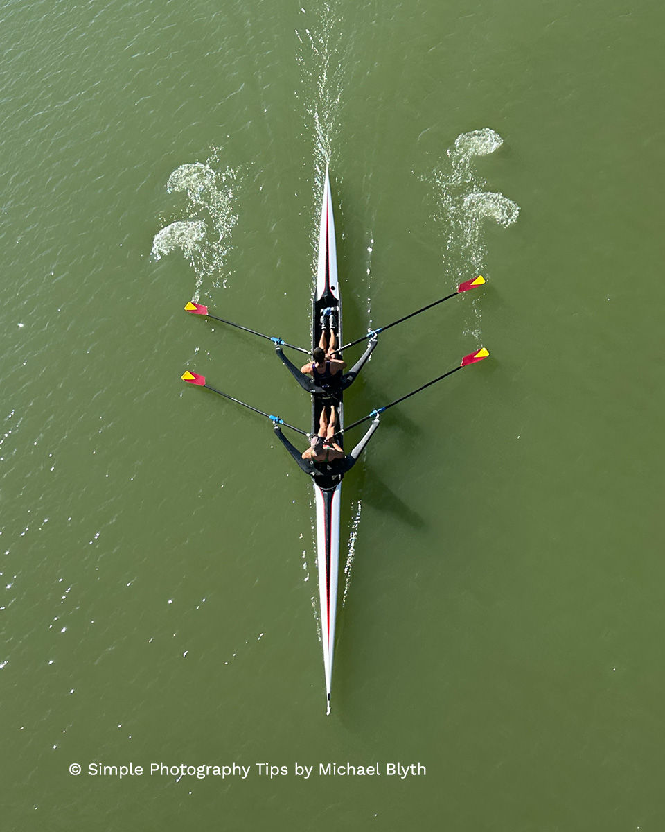 Two rowers paddle a skiff on green water. Oars splash symmetrically. Text reads: "© Simple Photography Tips by Michael Blyth."
