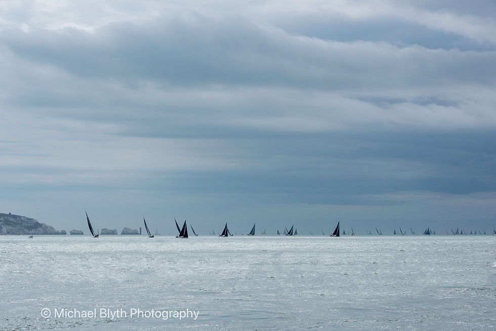 Admirals Cup 2025 Numerous sailboats with dark sails on a vast gray sea, under a cloudy sky. Text reads Michael Blyth Photography. Calm, serene mood.