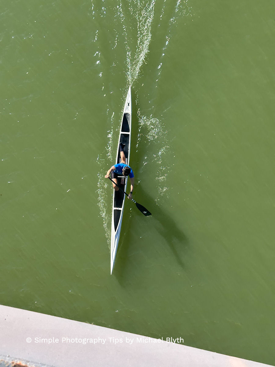 Aerial view of a person kayaking on green water, wearing a blue shirt. Ripples trail behind the kayak. Text: © Simple Photography Tips by Michael Blyth.