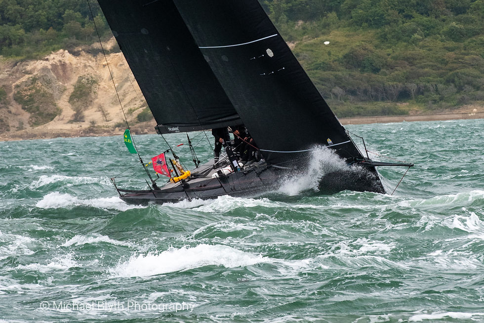 Sailboat with black sails cuts through rough green sea near a forested shore. rorc-admirals-cup-fastnet-yacht-race-2025