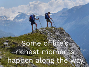 Two hikers standing on a grassy alpine ridge looking out across layered mountain peaks in the Vanoise region of the French Alps.