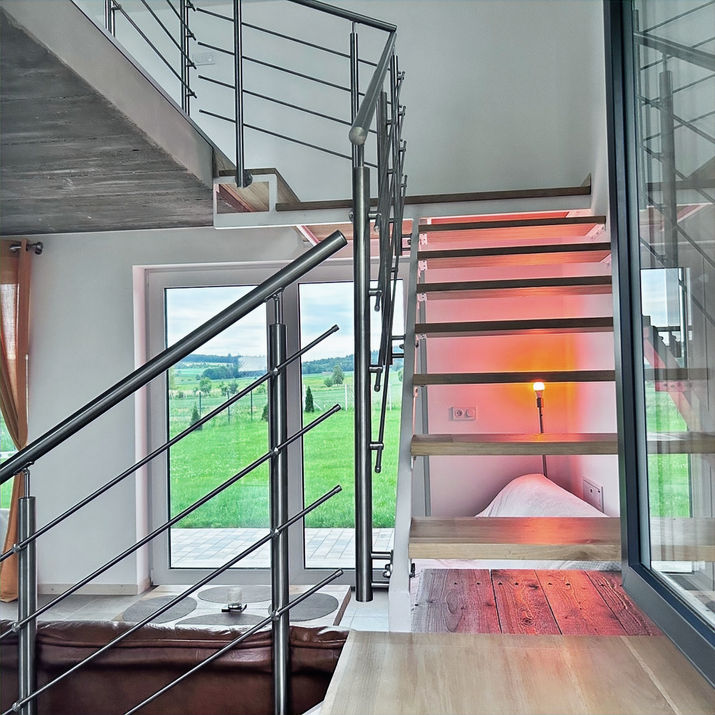 Dębowe schody w Nuka House z metalową balustradą, przy dużym oknie z widokiem na zielone pola.