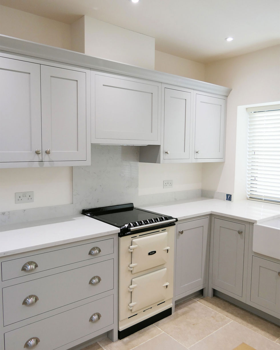 A corner of a kitchen showing light grey shaker style bespoke handcrafted cabinetry. There is a small age, a window with a closed blind and there is no kitchen equipment/utensils on show, it is very minimalistic.