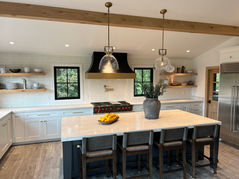 Modern farmhouse kitchen with white shaker cabinets and natural wood butcher-block island topped with industrial pendant lights. 

Professional gas range centered between black-framed windows. Open floating wood shelves on white walls. Stainless steel appliances and warm oak hardwood flooring. Light wood ceiling beam adds rustic warmth to the bright, open-concept design.