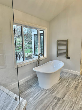 Contemporary master bathroom featuring a freestanding white soaking tub centered beneath a picture window. Gray wood-look tile flooring with linear grain pattern flows throughout. Walk-in glass shower enclosure with white subway tile. Chrome towel warmer mounted on pale gray walls. Natural daylight illuminates the clean, minimalist design.