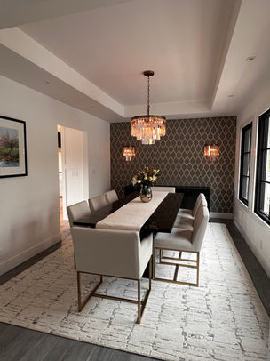 11:41 AMTransitional dining room with navy geometric quatrefoil wallpaper accent wall and white tray ceiling. Modern rectangular dining table with gray upholstered chairs and clear acrylic ghost chairs. Three-tiered crystal chandelier centerpiece flanked by matching wall sconces. Dark hardwood flooring with light geometric area rug. Black-framed windows provide natural light in the long, gallery-style space with white wainscoting.