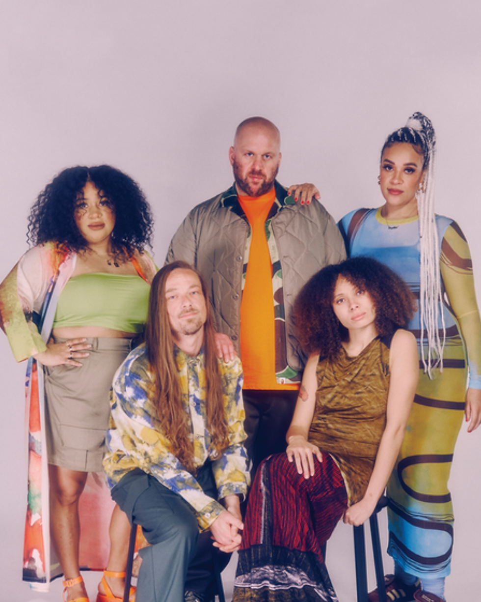 Wooden saunas on a deck at London sunset with skyscrapers in the background. Warm golden light, rustic barrels, greenery, and a water fountain with text: "DRINK."