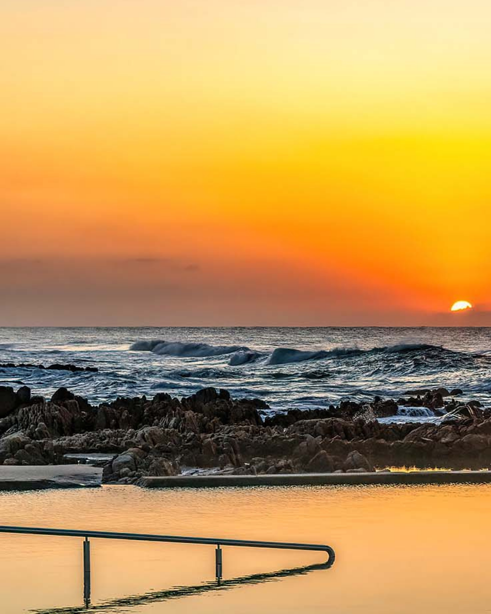 Sunset over a rocky shore with gentle waves, vibrant orange sky, and a reflective tidal pool in the foreground with a railing. Peaceful mood.