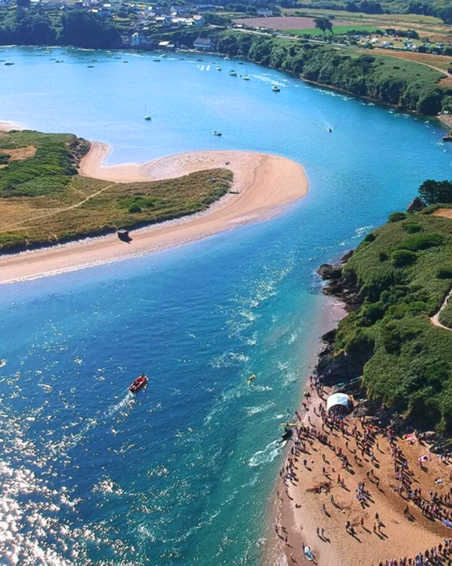Aerial view of a sunny beach scene with a crowd of swimmers on the sand, a boat in blue water, lush green hills, and boats anchored in a distant bay.